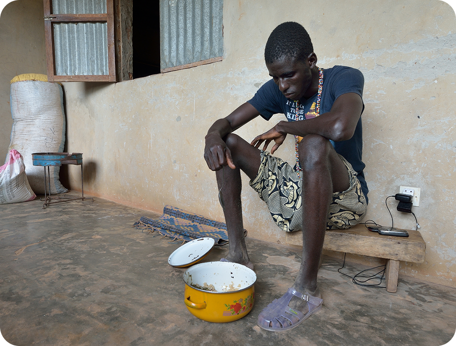 A man in a humble indoor setting sits on a bench beside a pot of food, appearing contemplative, highlighting themes of poverty and resilience.