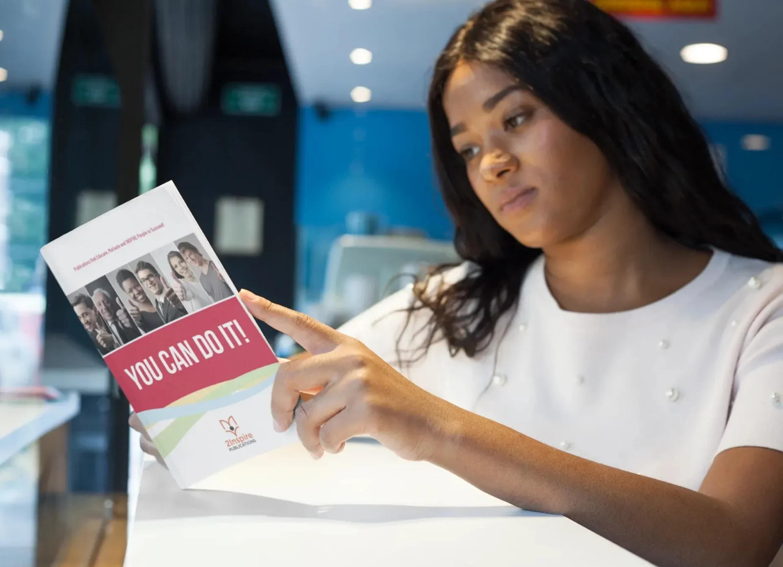 A confident young woman in a white top reads a motivational booklet titled “You Can Do It!” by 2Inspire Publications in a modern indoor setting.